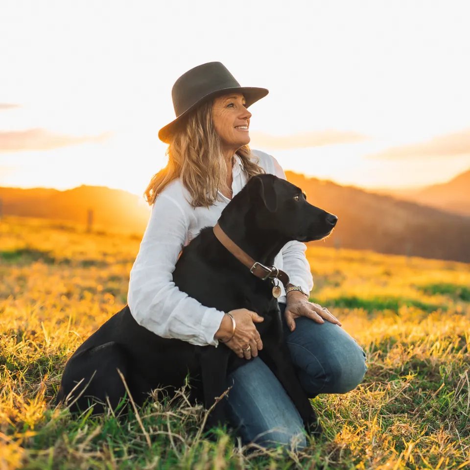 Cyndi kneeling in a sunlit paddock with her black dog
